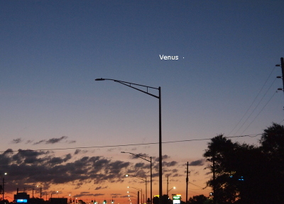 [This is a street view with the street lights still lit while the lower portion of the sky starts to turn red-orange. There is a line of clouds on the horizon below which is the orange-red sky and above which is the clear dark blue sky. Venus is a bright white spot above and to the right of a street light post. To the left of Venus is its name in white letters.]